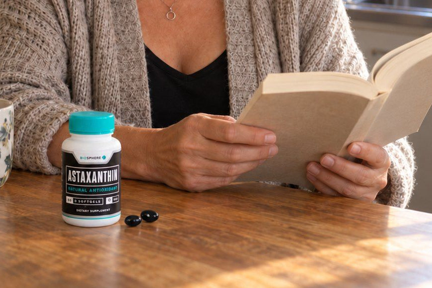 Hands holding an open book at a kitchen table, with a bottle of Biosphere Astaxanthin and two black softgels in front