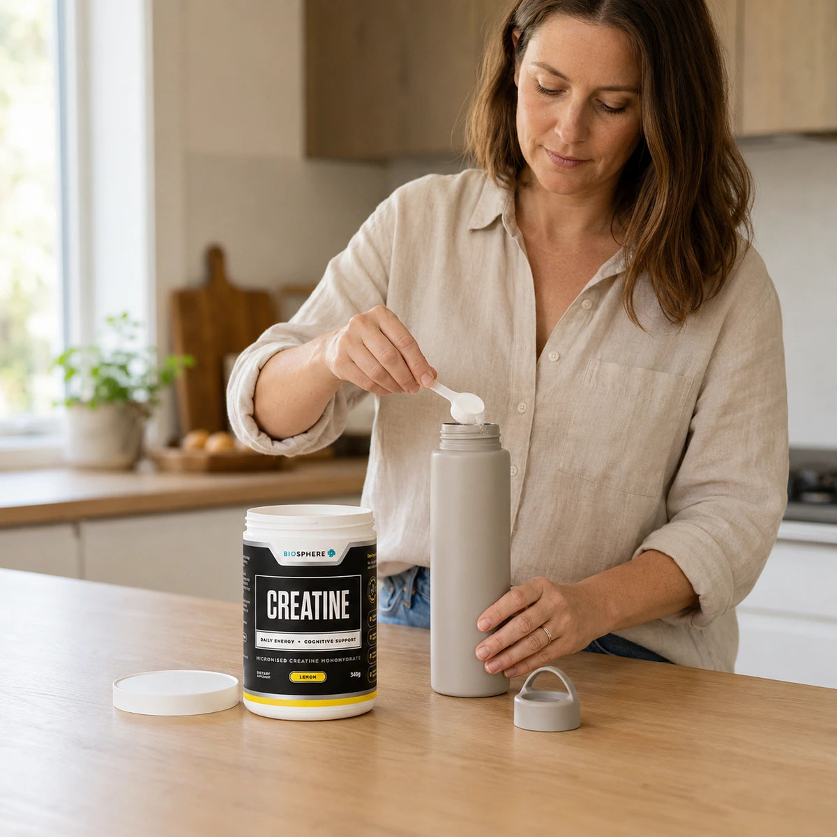 Woman adding a scoop of Biosphere Creatine to a drink bottle, tub on the bench